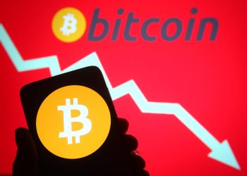 A hand holding up a phone with bitcoin on it. A white arrow points downward on a red background