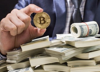 Picture of a hand holding a bitcoin over a pile of cash